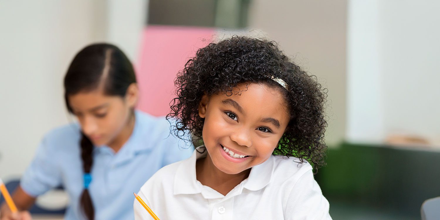 Smiling student holding a pencil while a classmate writes nearby.